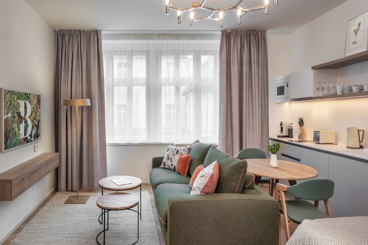 A modern living room with a green sofa, a round wooden table, and a kitchen area with a microwave and toaster.