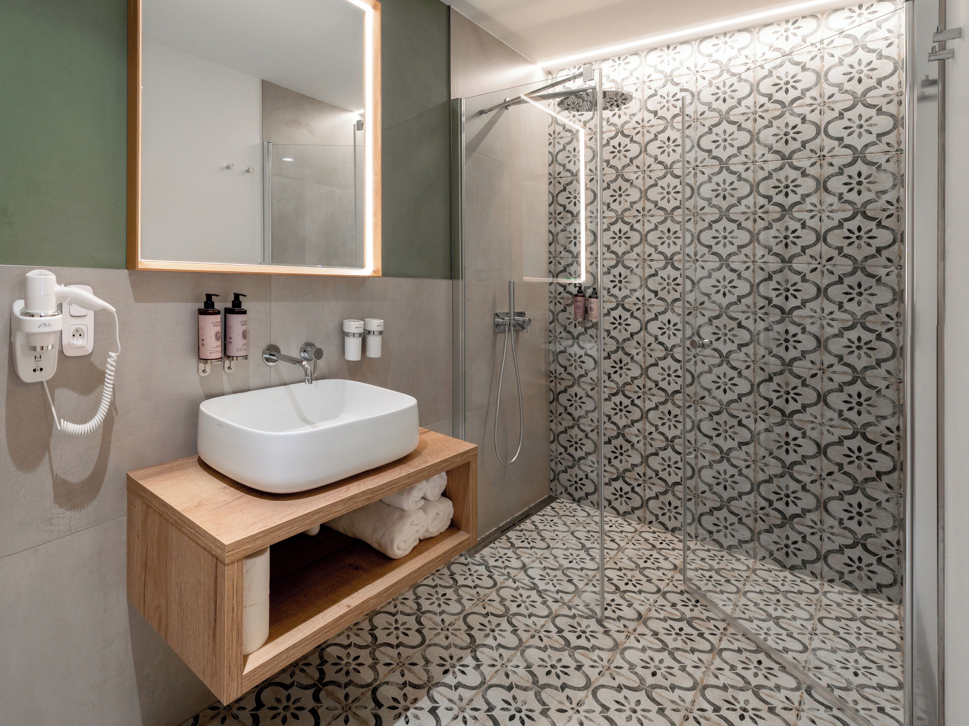 Un baño moderno con lavamanos blanco, tocador de madera y zona de ducha con azulejos estampados.