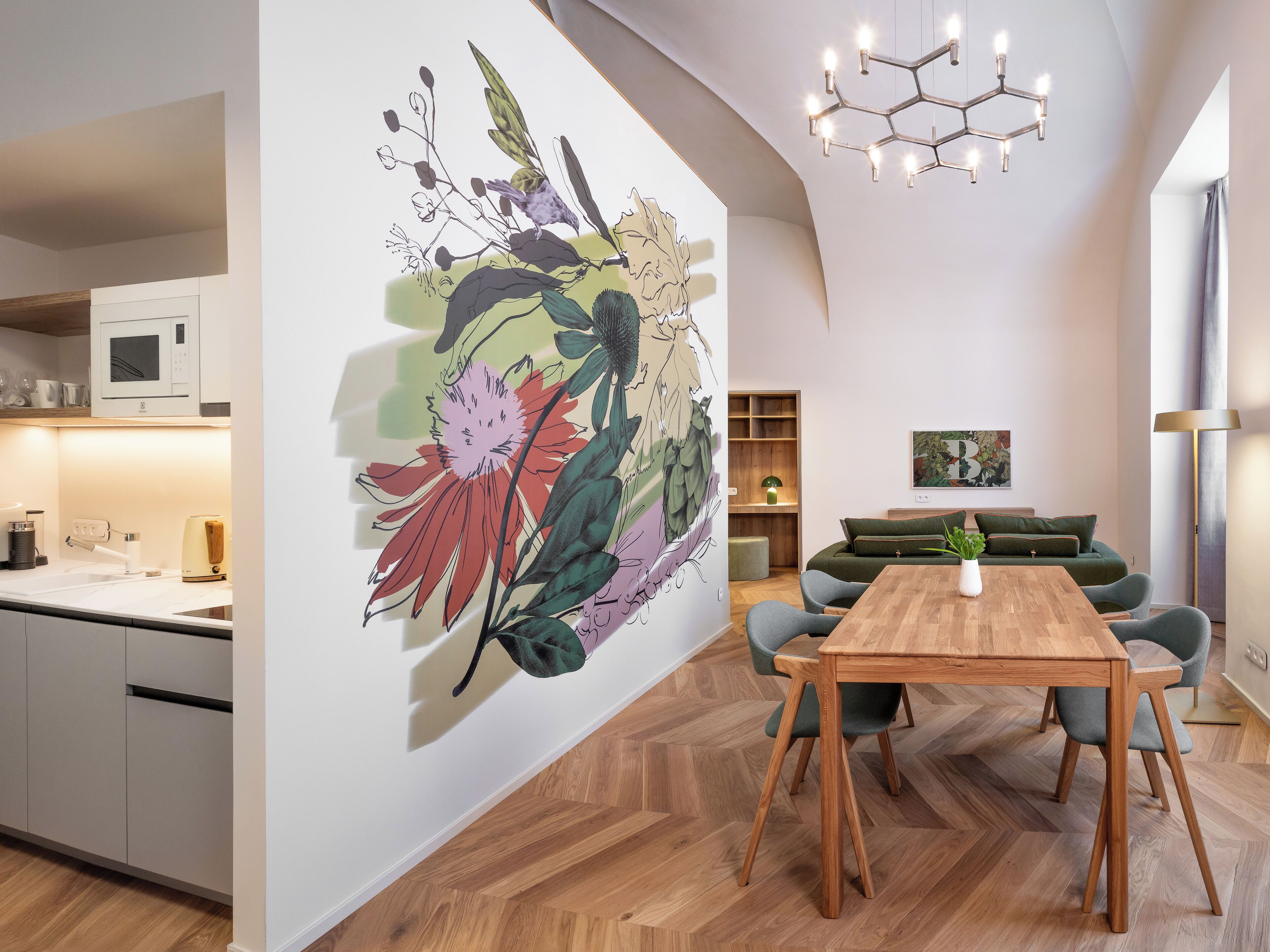 A modern kitchen with a microwave, a coffee machine, and a sink. There is a dining area with a wooden table and chairs, and a green sofa. A large floral mural is on the wall, and a chandelier hangs from the ceiling.