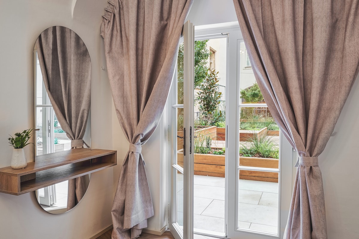 A room with a wooden floor, a round mirror, a small shelf with a plant, and a view of a garden through a glass door.
