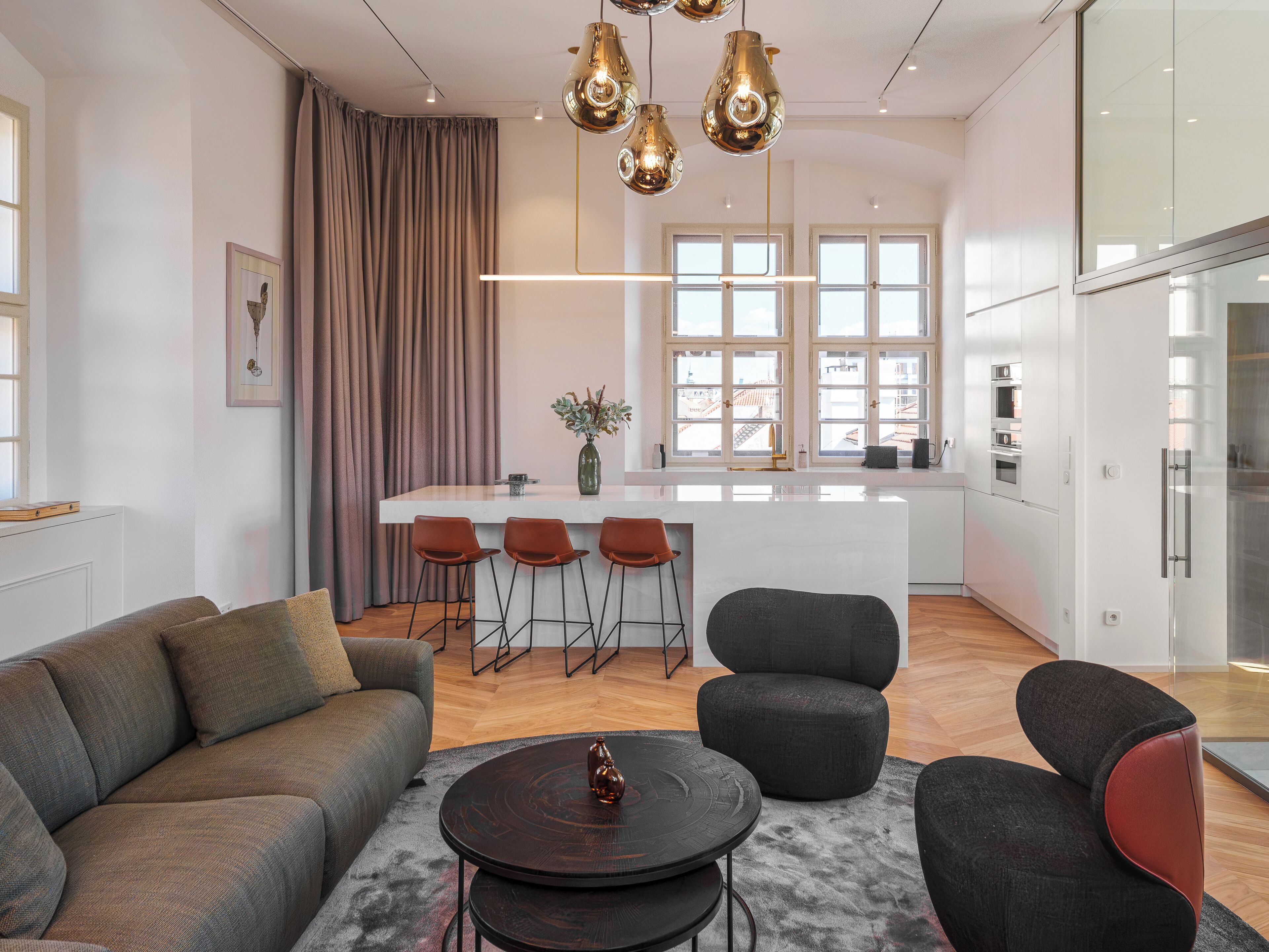 Una sala de estar moderna con sofá, sillas, una mesa de centro y una cocina con barra y ventanas amplias.