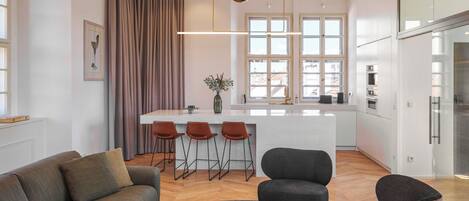 Una sala de estar moderna con sofá, sillas, una mesa de centro y una cocina con barra y ventanas amplias.