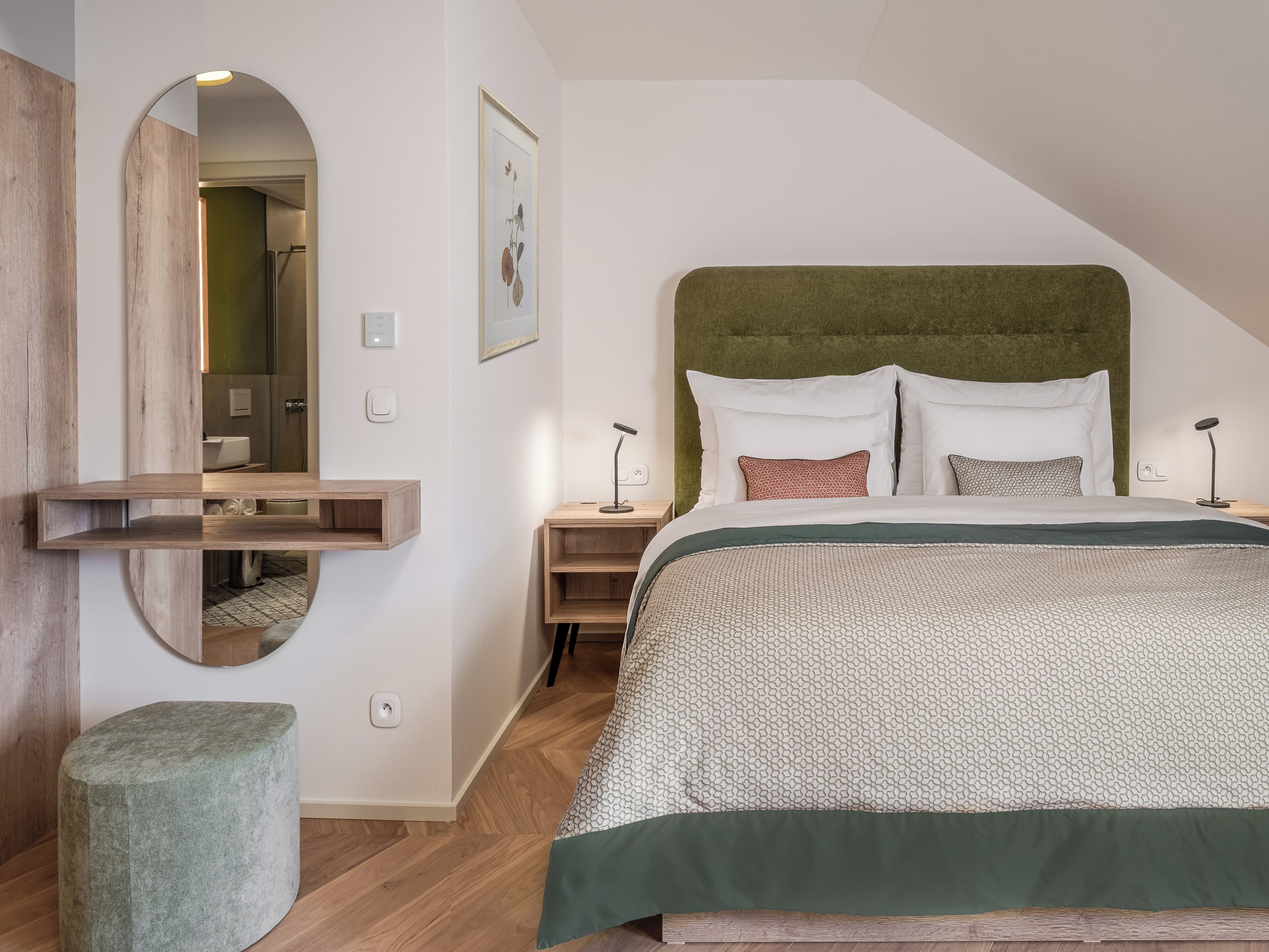 A bedroom with a bed, a bedside table, a nightstand, a mirror, and a green ottoman.