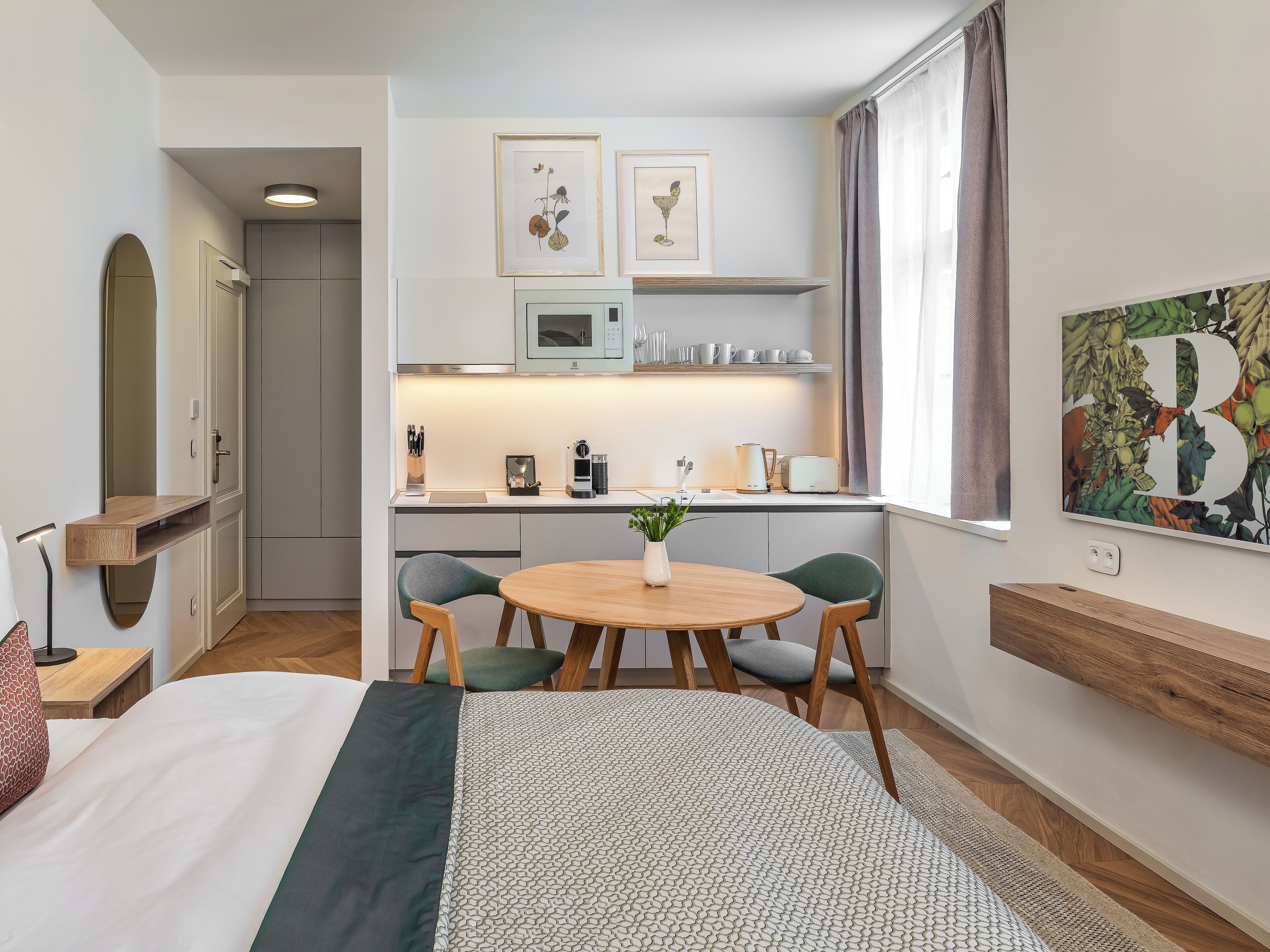 A modern hotel room with a bed, a round wooden table with two chairs, a small kitchen area with a microwave, and a large window with curtains.