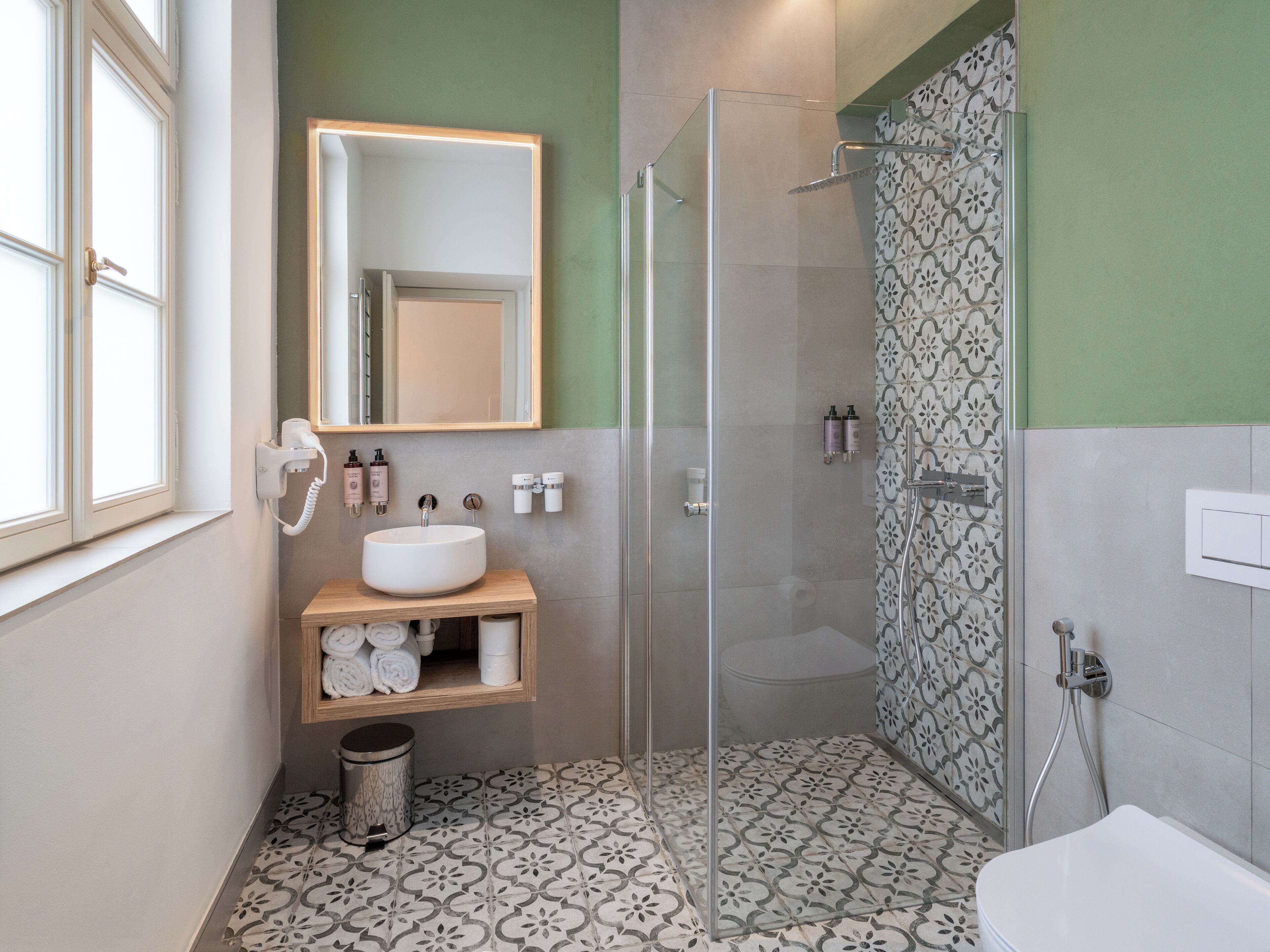 A bathroom with a glass-enclosed shower, a sink with toiletries, and patterned floor tiles.