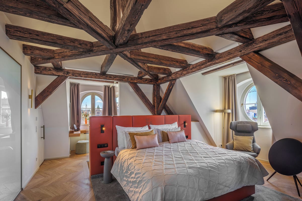 A bedroom with a sloped ceiling, exposed wooden beams, a red headboard, a large bed with multiple pillows, a round chair, and a view through an arched window.