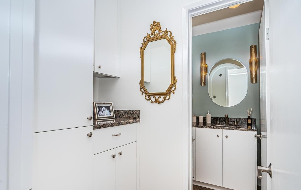 Une salle de bain avec des meubles blancs, un miroir encadré d’or et un comptoir en granit.