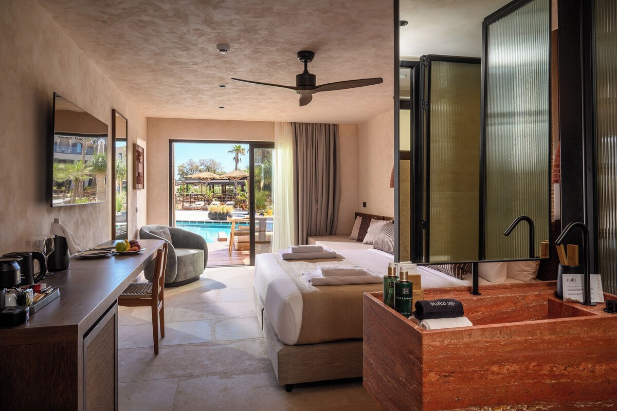 A hotel room with a bed, a desk, a chair, a bathroom, and a view of an outdoor pool area.