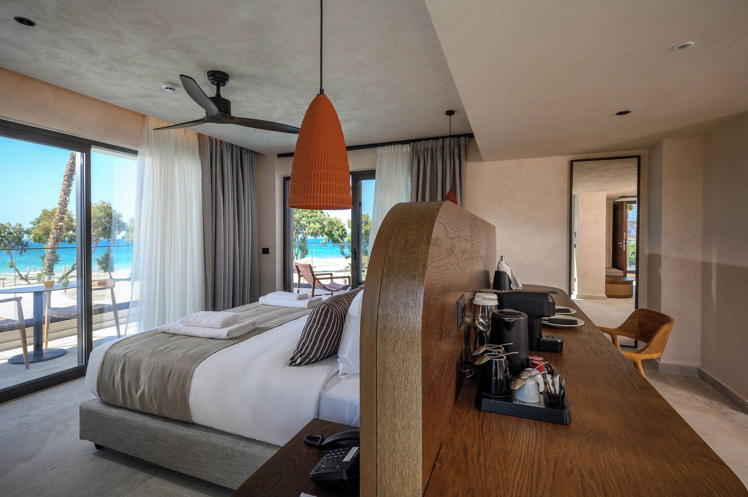 A modern hotel room with a large bed, a wooden desk with amenities, a ceiling fan, and a view of the beach.