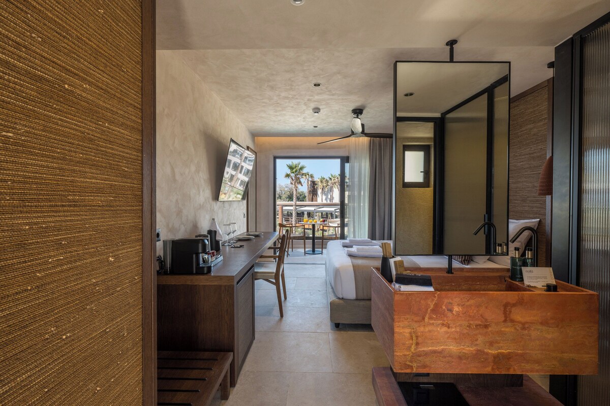 A modern hotel room with a kitchen area, a dining table, a sofa, and a large window with a view of palm trees.