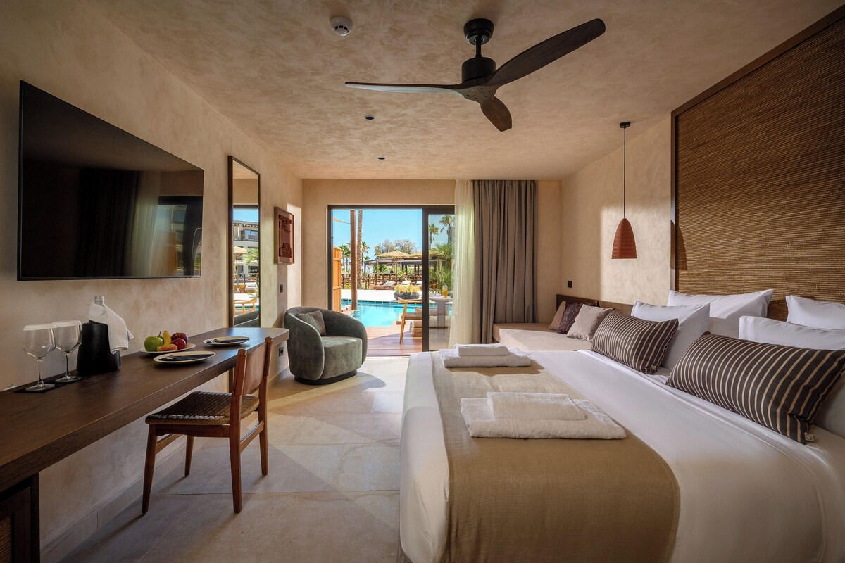 A hotel room with a bed, a desk, a chair, a TV, and a view of an outdoor pool area.