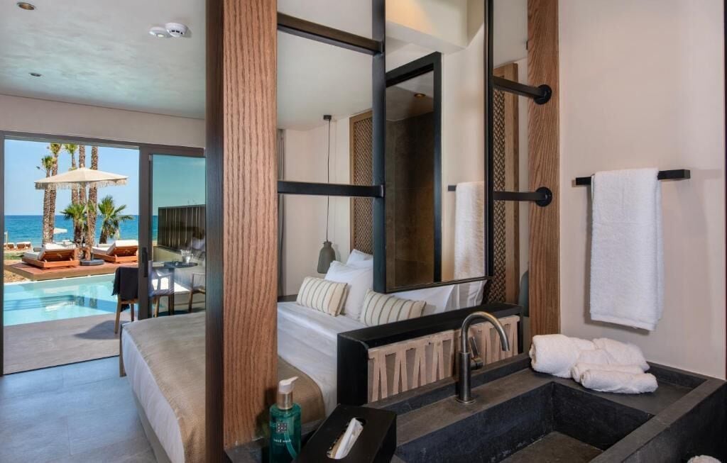 A modern hotel room with a large bed, a bathroom with a black sink, and a view of a pool and palm trees.
