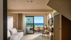 Ein Hotelzimmer mit einer Couch, einem Esstisch mit Stühlen, einem Schreibtisch mit einem Computer und einem großen Fenster mit Blick auf den Strand und den Ozean.