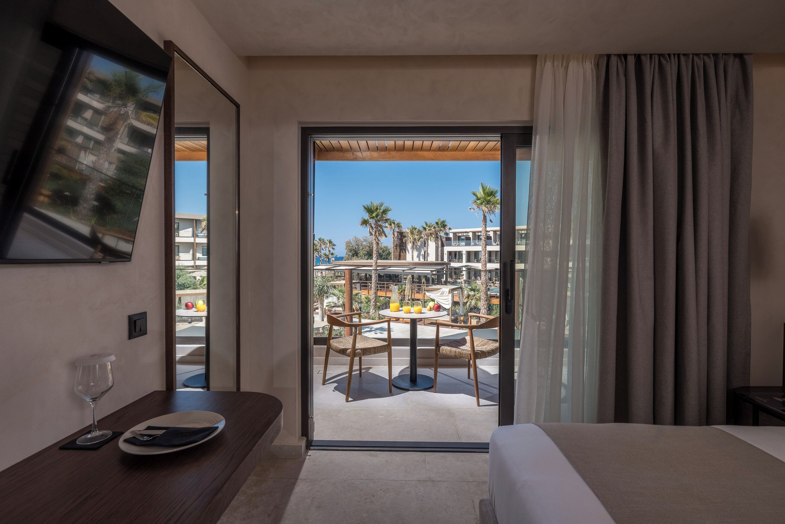 Habitación de hotel con cama, mesa de comedor con sillas, televisor montado en la pared y vista al área de la piscina del resort.