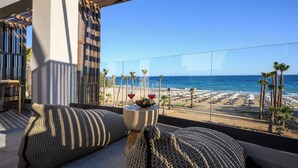 Ein Balkon mit Blick auf den Strand, ein Tisch mit zwei Gläsern Rotwein und ein gewebter Sitzhocker.