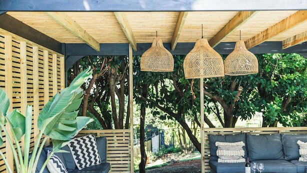 Un espace extérieur aménagé, abrité et doté de poutres en bois, d’un canapé, d’une table et de lampes en rotin suspendues.