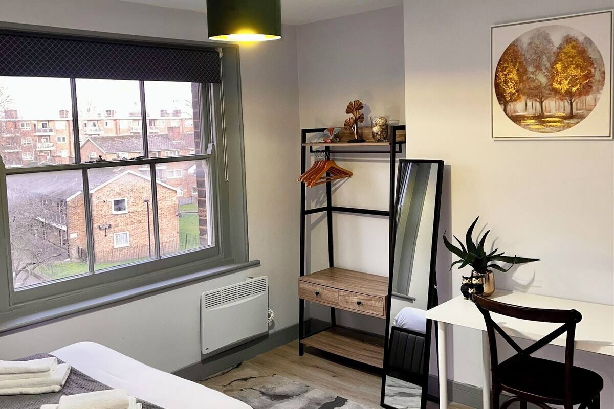 A modern bedroom with a bed, a desk, a chair, a radiator, a window with a view of buildings, and a hanging light fixture.