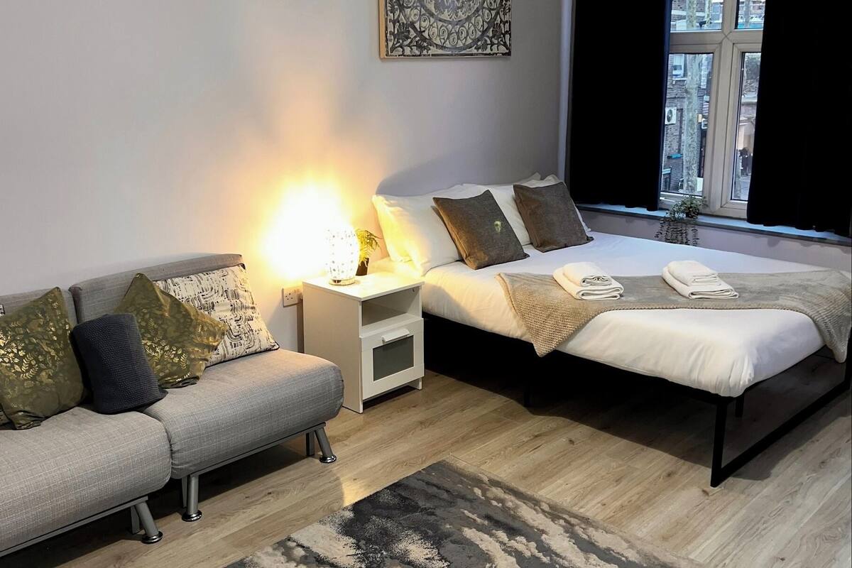 A modern bedroom with a sofa, a bed, a nightstand with a lamp, and a large window with curtains.