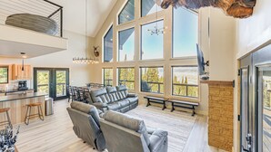 Ein geräumiger Wohnbereich mit einem großen, grauen Ecksofa, einem Kamin aus Stein und einer hohen Decke mit Dachfenstern.