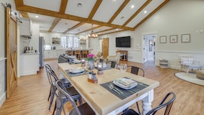 Une cuisine spacieuse avec des poutres en bois, un coin repas avec une longue table dressée pour six personnes, et un salon avec un canapé et une télévision.