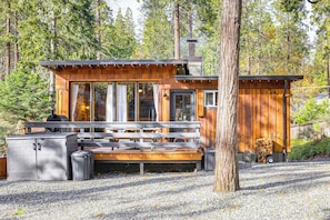 A cabin with a deck, a grill, and a large window overlooking a forest.