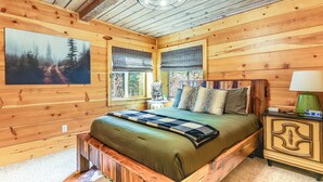 A wooden cabin bedroom with a bed, a nightstand, a lamp, and a painting of a forest on the wall. - Walk to Golf, Lake: Twain Harte Cabin w/ Fireplace (Twain Harte)