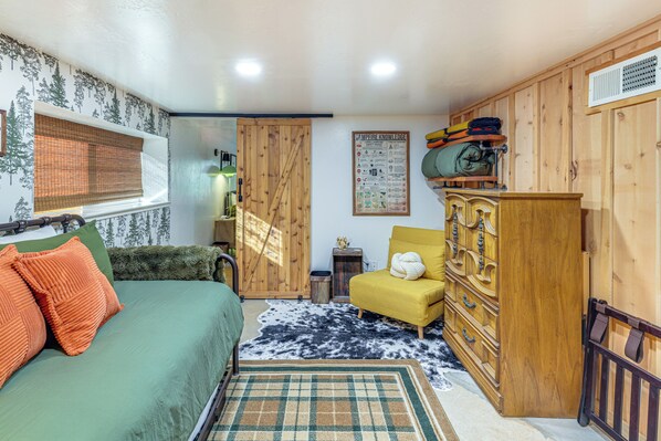 A cozy room with a bed, a yellow armchair, a wooden dresser, and a window with blinds.