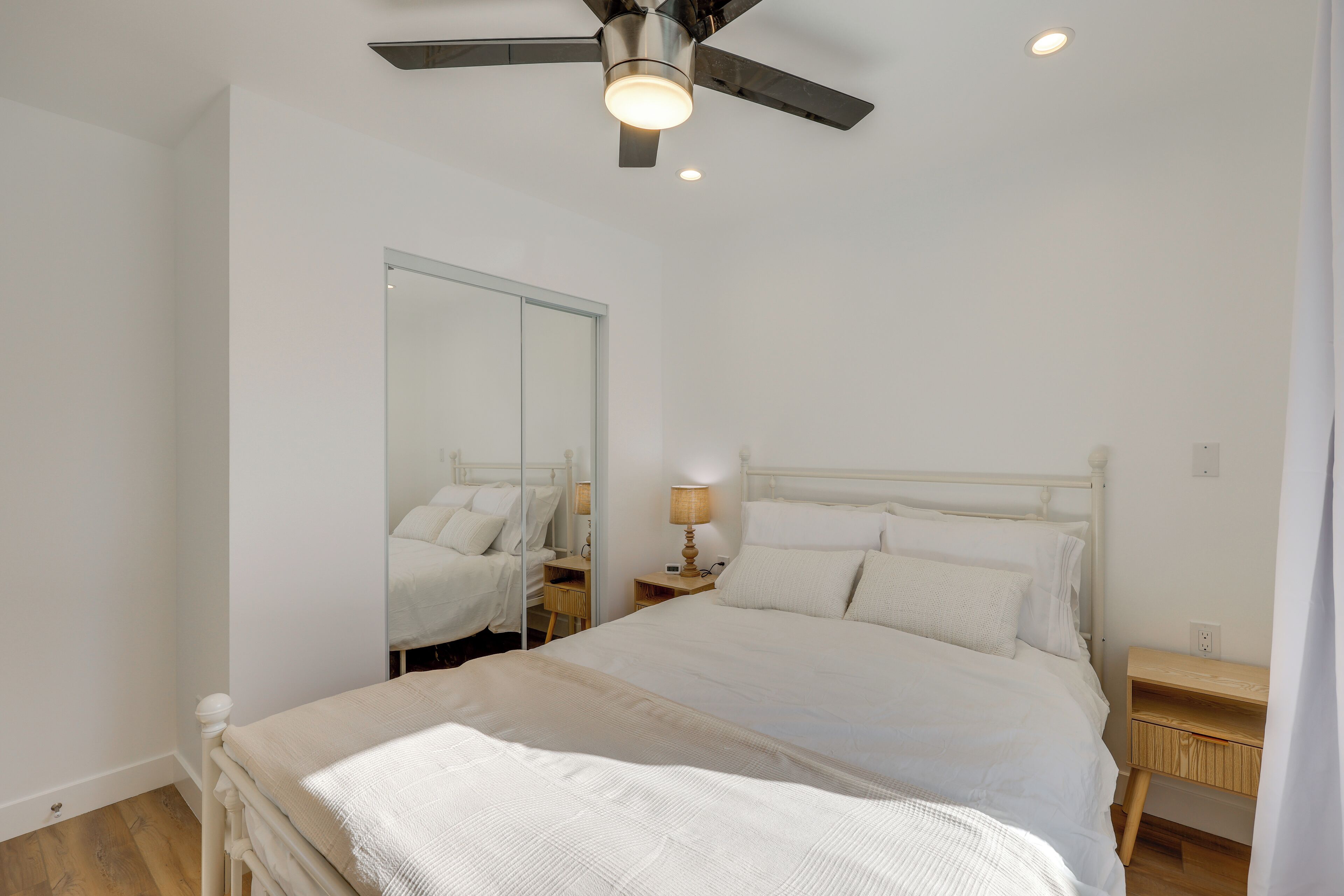 A bedroom with a bed, a ceiling fan, a bedside table with a lamp, and a mirror.
