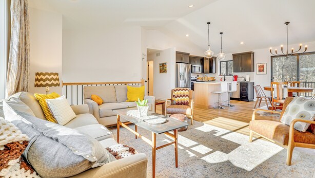 A spacious living area with a sofa, armchairs, a coffee table, and a dining table. A kitchen is visible in the background with modern appliances and a large window. - Deck w/ Forest Views: Modern Home in Rochester! (Rochester)