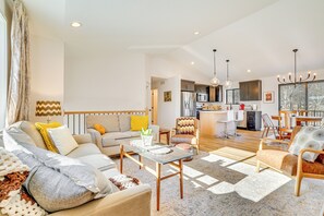 A spacious living area with a sofa, armchairs, a coffee table, and a dining table. A kitchen is visible in the background with modern appliances and a large window. - Deck w/ Forest Views: Modern Home in Rochester! (Rochester)