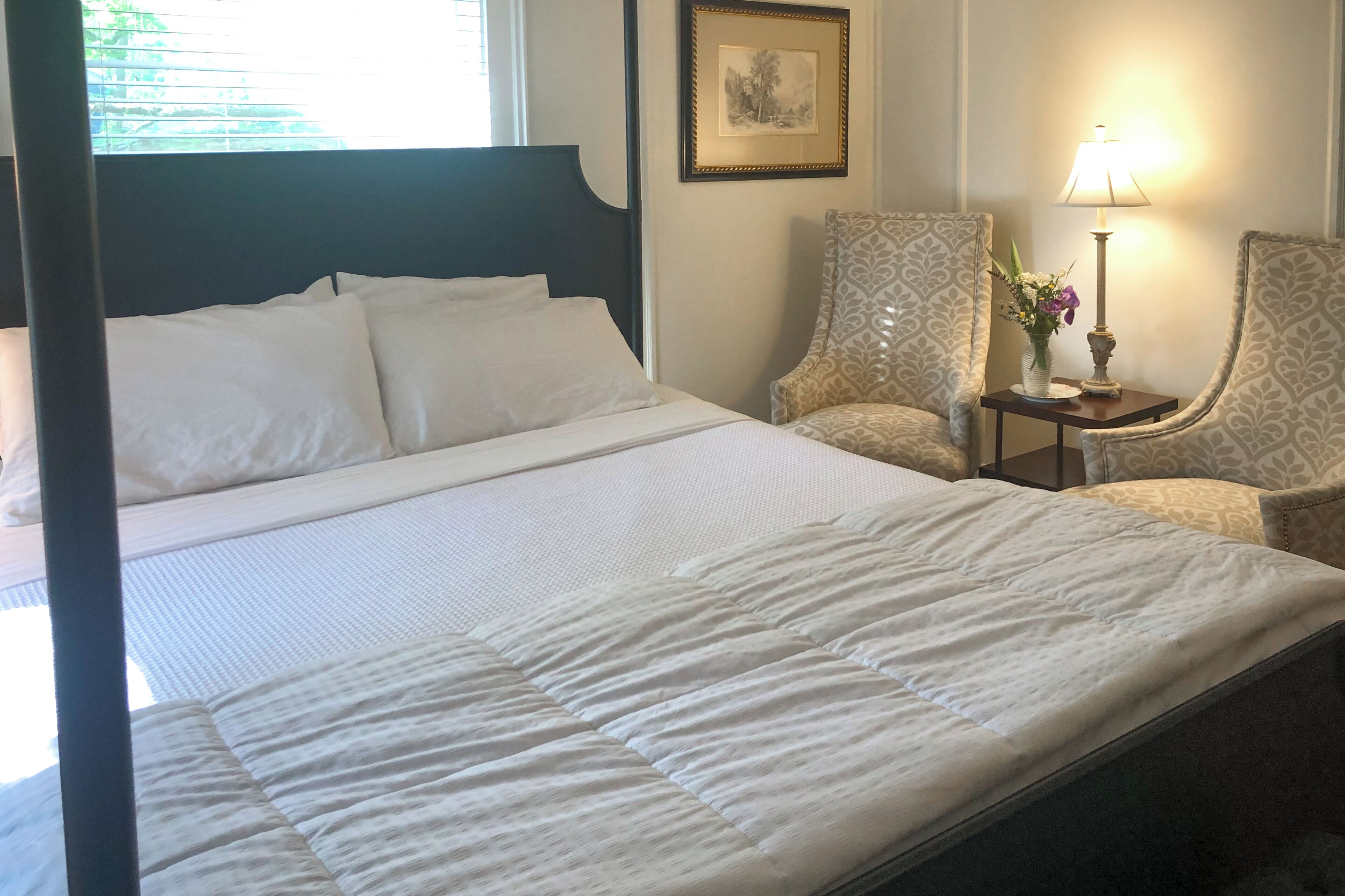 A neatly made bed with white linens, a bedside table with a lamp and vase, and patterned armchairs.