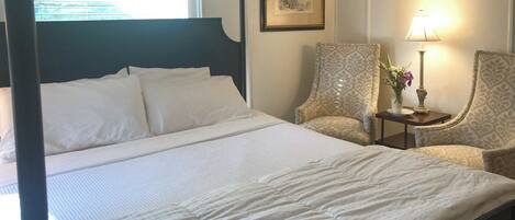 A neatly made bed with white linens, a bedside table with a lamp and vase, and patterned armchairs.