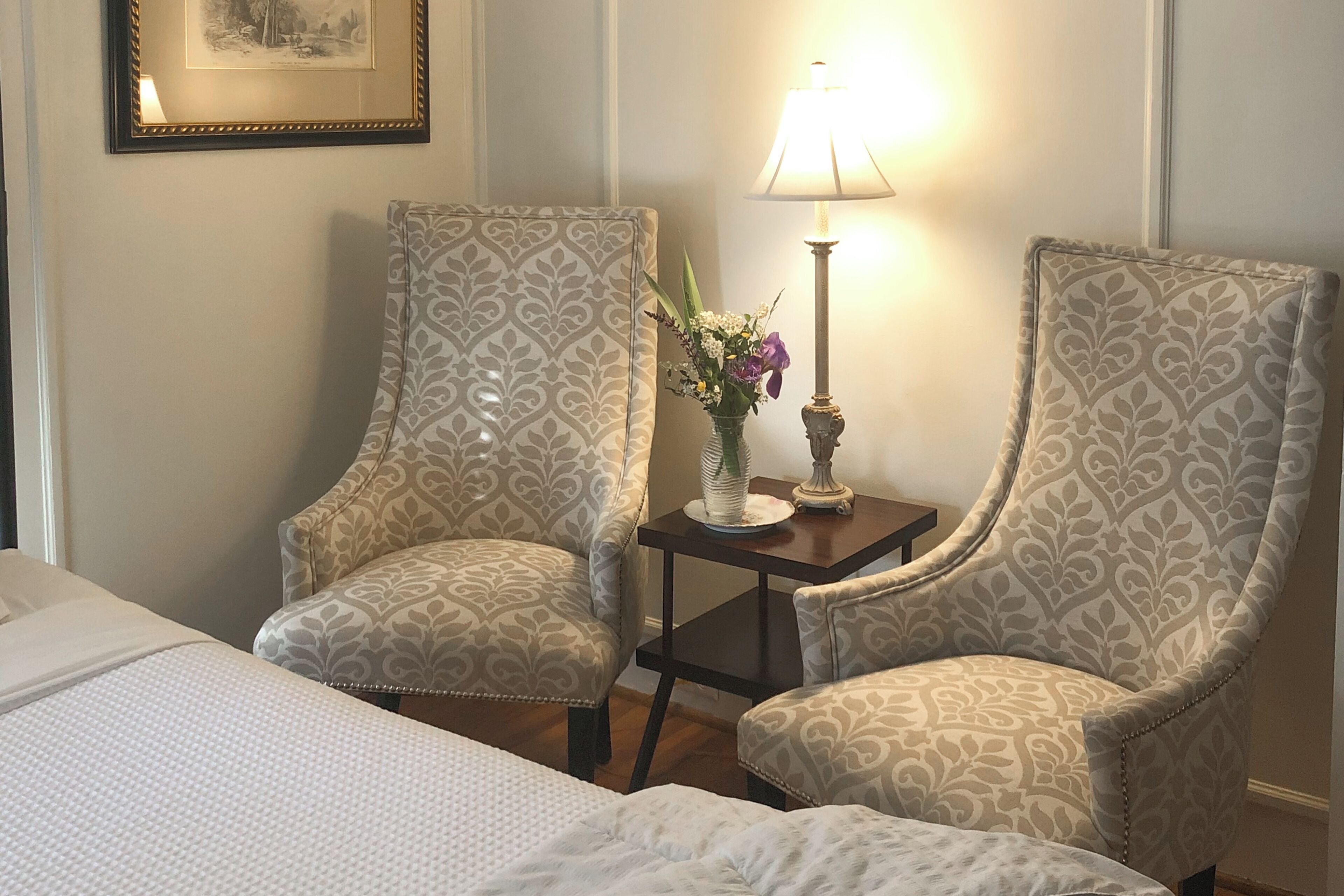 Een hotelkamer met twee gestoffeerde fauteuils, een tafeltje met een lamp en een bloemenvaas, en een bed met een wit dekbed.