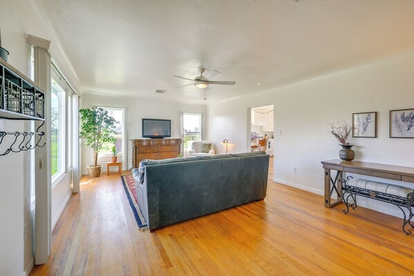 A spacious living room with hardwood flooring, a large grey sofa, a ceiling fan, and a television on the wall.