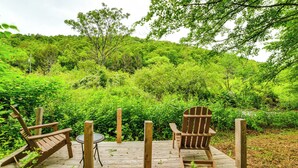 Una terraza de madera con dos sillas Adirondack y una mesita, rodeada de abundante vegetación y árboles.