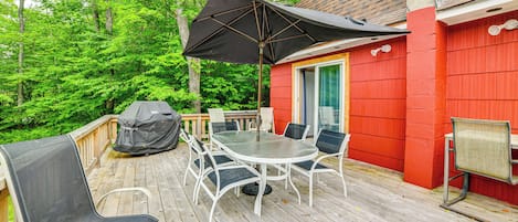 A wooden deck with a dining table and chairs, a grill, and a red building in the background.