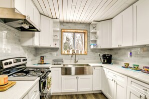 Una cocina con armarios blancos, un lavamanos de acero inoxidable, una estufa y una ventana sobre el lavamanos.