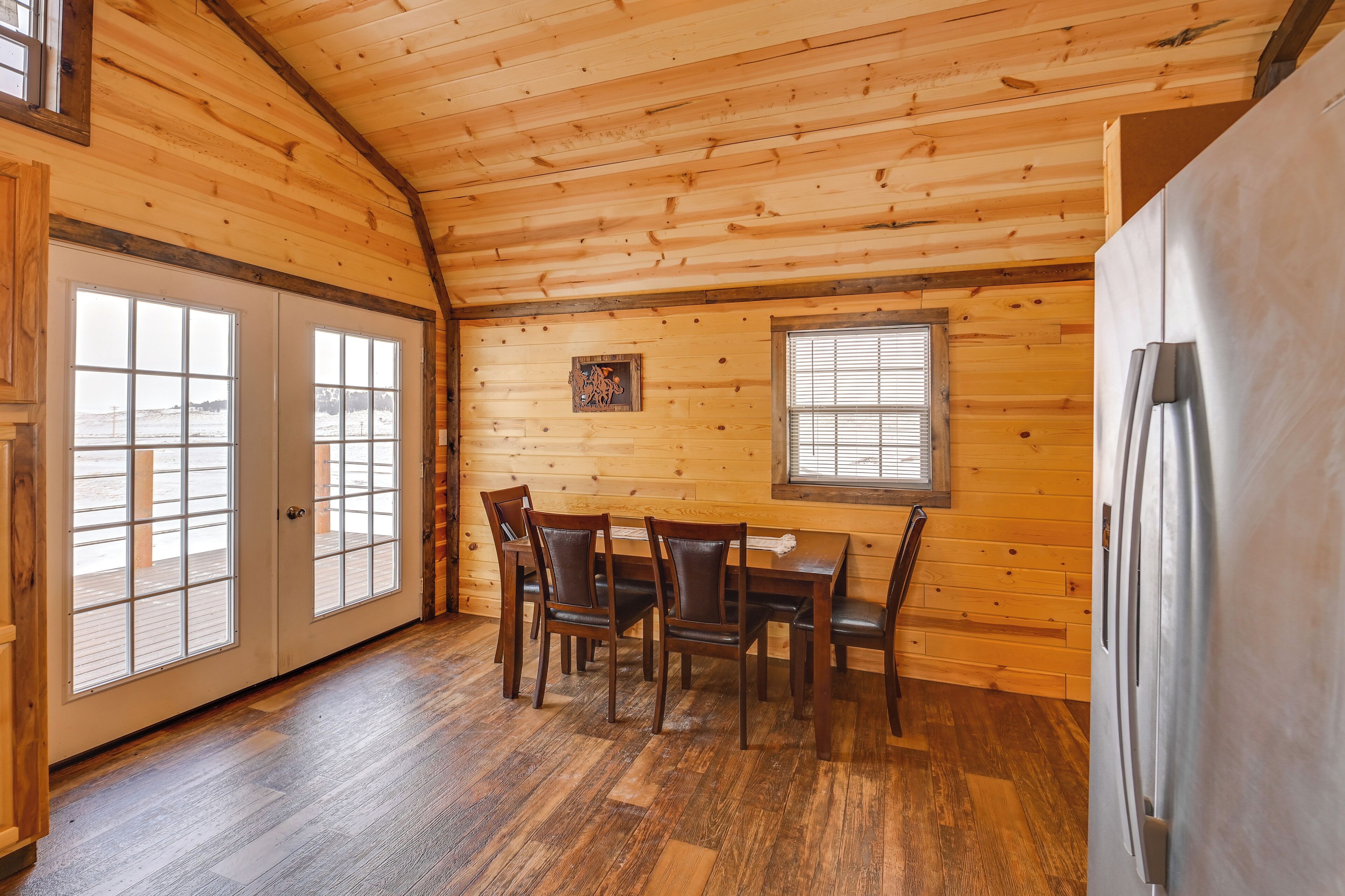 Un interior de madera con una mesa de comedor, sillas, un refrigerador y ventanas grandes.