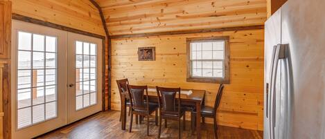 A wooden interior with a dining table and chairs, a refrigerator, and large windows.