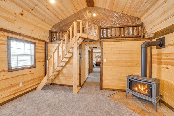 Interior de una cabaña de madera con una escalera central, chimenea y ventana.