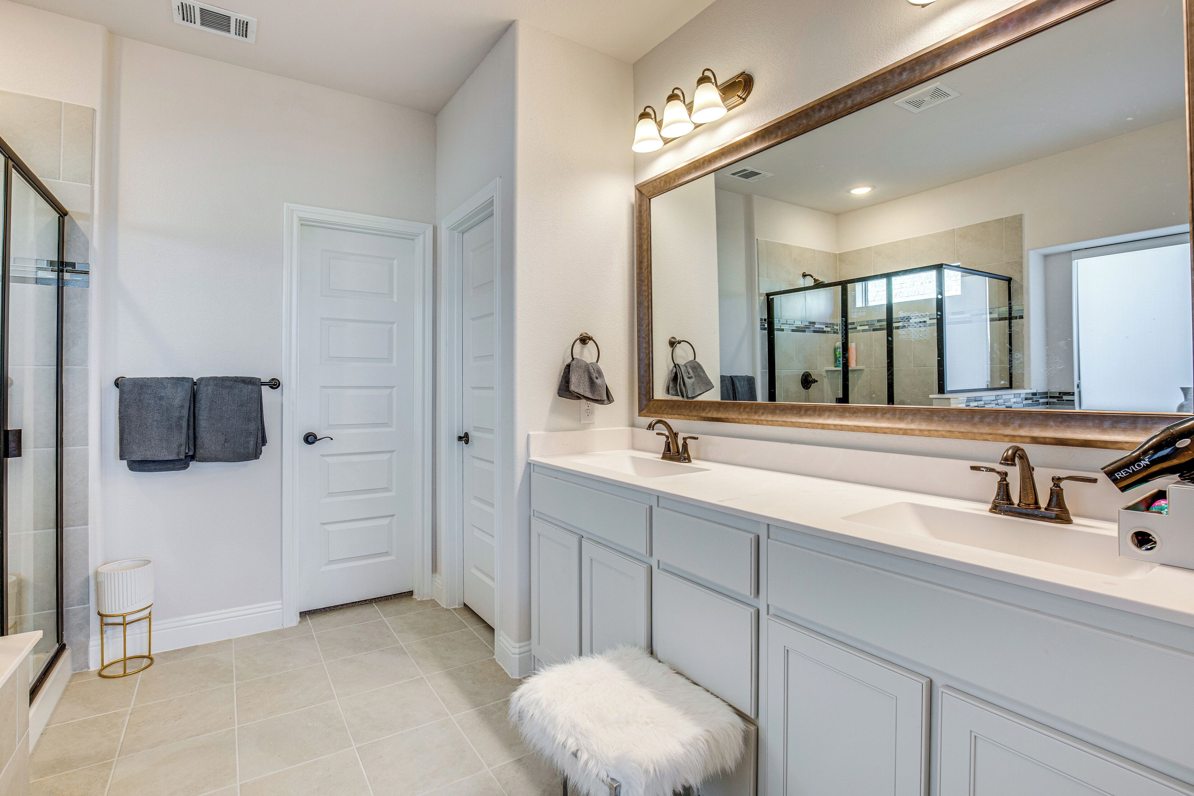 A bathroom with a large mirror, white cabinets, a white sink, and a fluffy white stool.