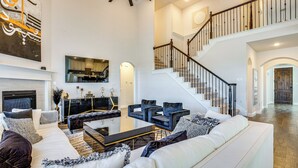 A spacious living room with a white sofa, black and white patterned throw pillows, a black coffee table, and a fireplace. A staircase with a wooden railing leads to an upper level. - 6 Mi to Lake Lewisville: Chic Retreat in Frisco! (Frisco)