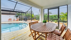 Ein Poolbereich mit Holztisch und -stühlen, einem kleinen Topfpflanzen und Blick auf ein Haus und Grünanlage durch große Fenster.