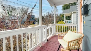 A porch with a wicker chair and a white railing, overlooking a residential area with houses and trees. - 1 Mi to Dtwn: Home w/ Backyard in Raleigh! (Raleigh)