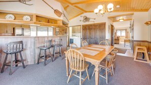 A wooden dining area with a long table and several chairs, a bar area with stools, and a kitchen visible in the background.