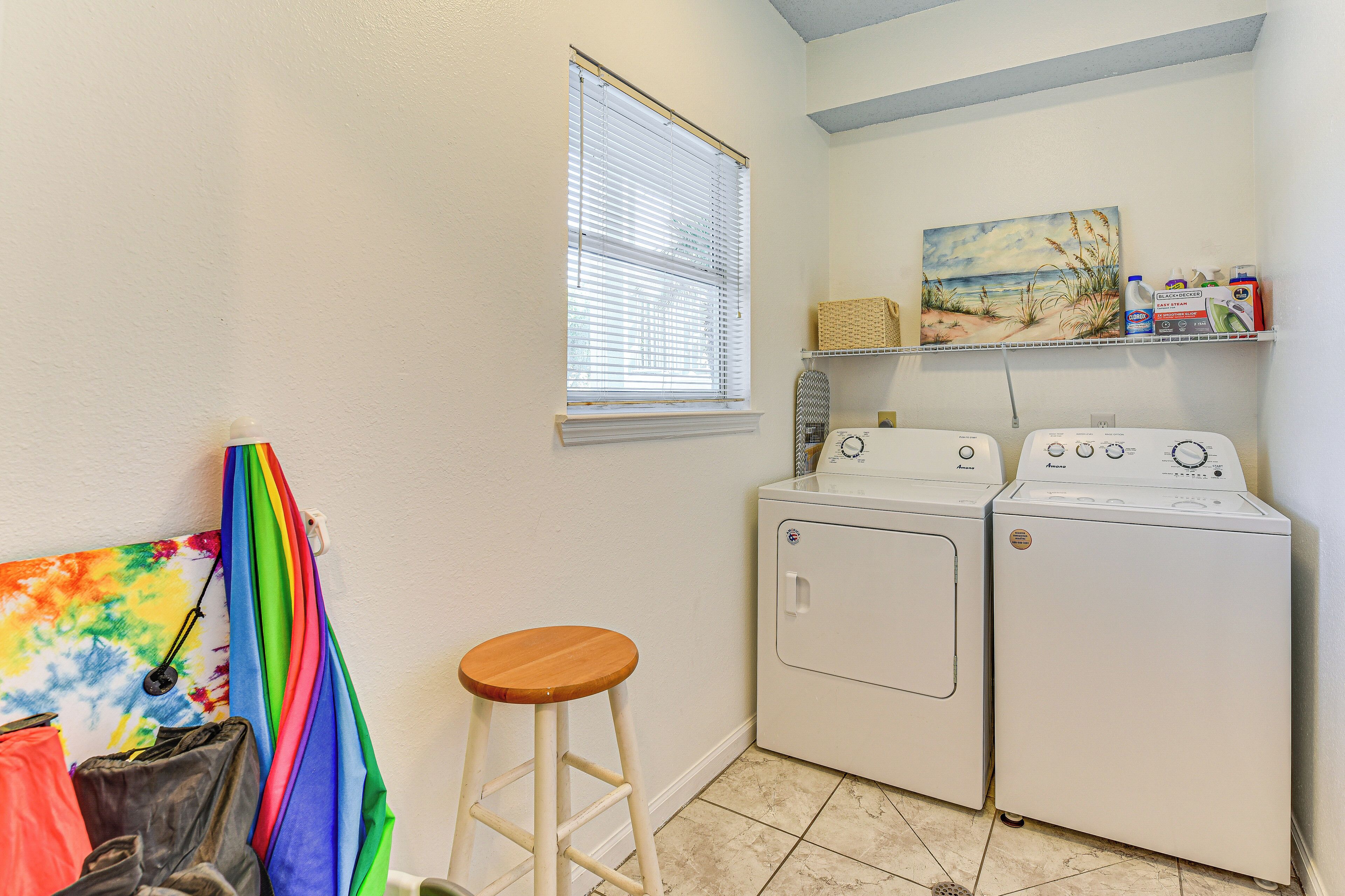 A laundry room with two white washing machines, a wooden stool, a window with blinds, and a shelf with cleaning supplies.