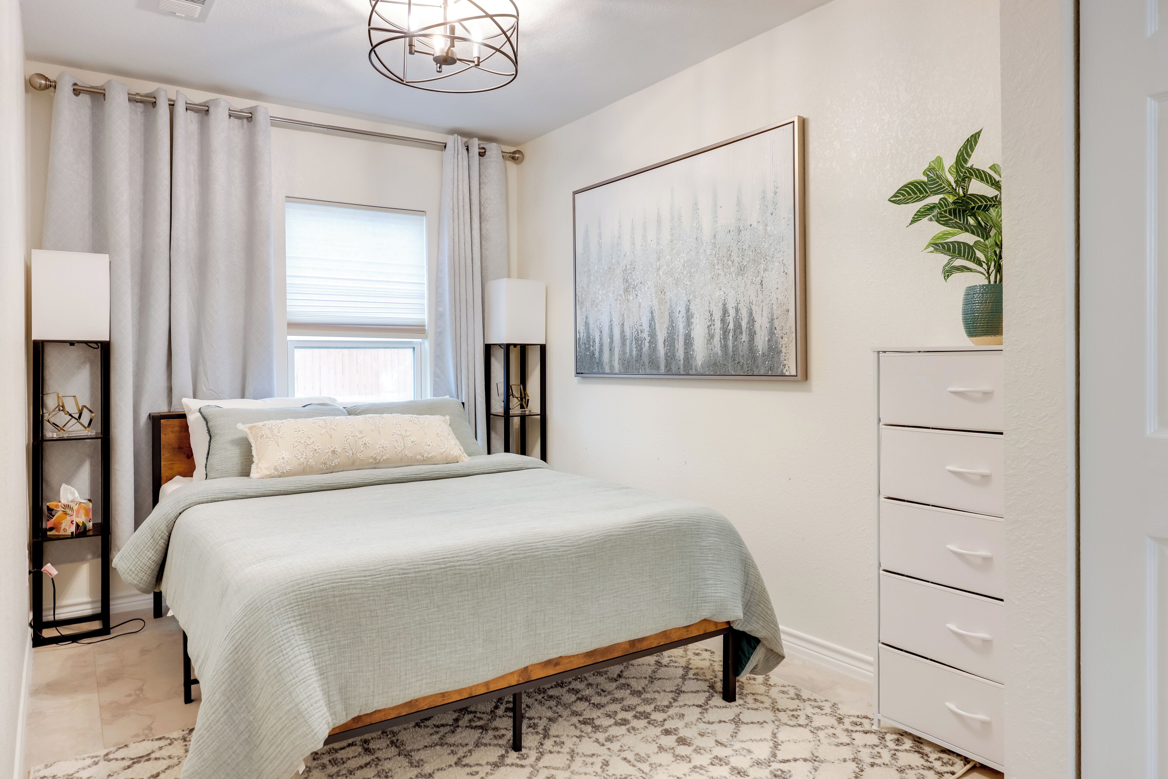 A bedroom with a bed, a nightstand, a lamp, a dresser, a window with curtains, a painting, and a plant.