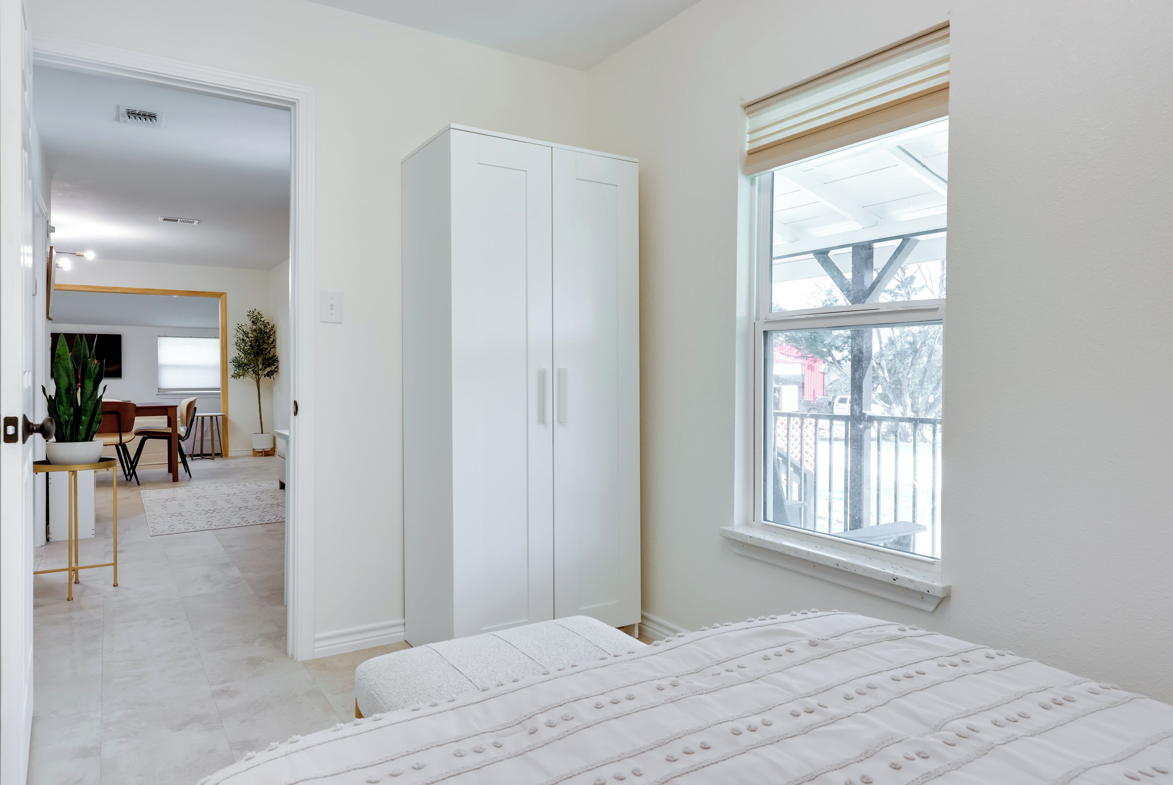 A bedroom with a bed, a wardrobe, a window with blinds, and a view of the outside.