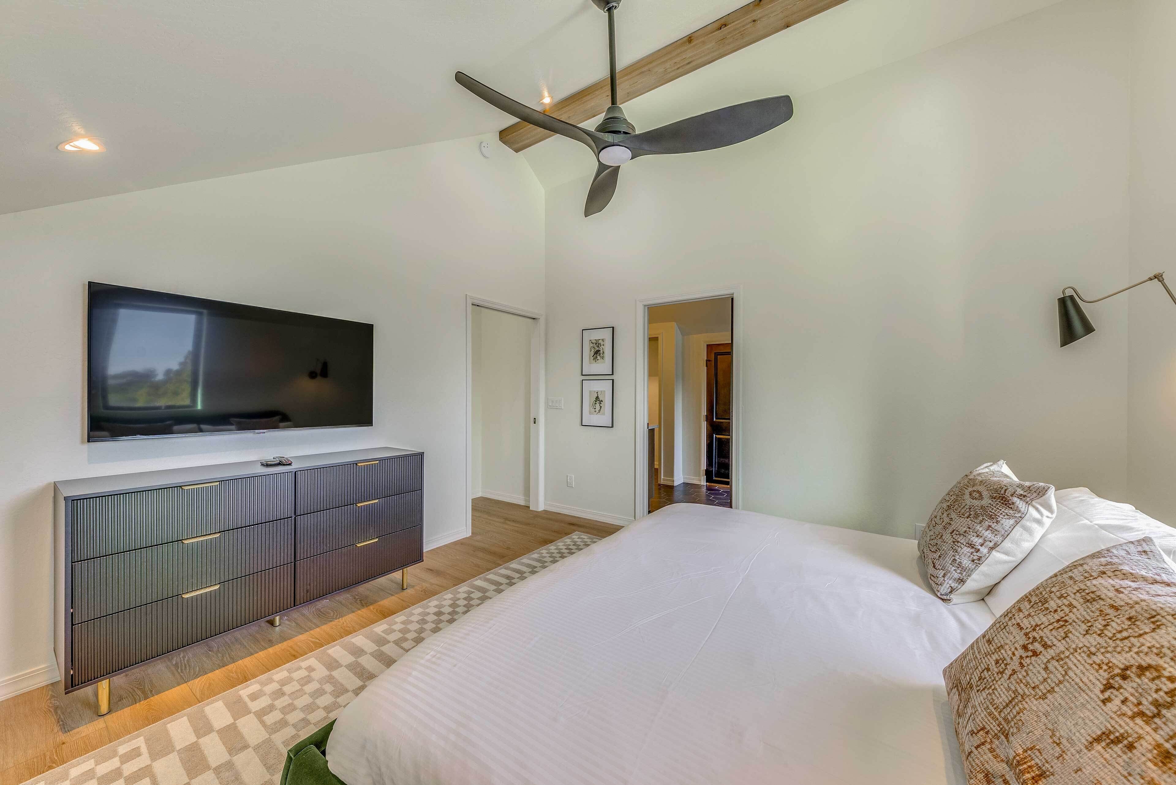 A bedroom with a bed, a TV mounted on a cabinet, a ceiling fan, and a hallway leading to another room.