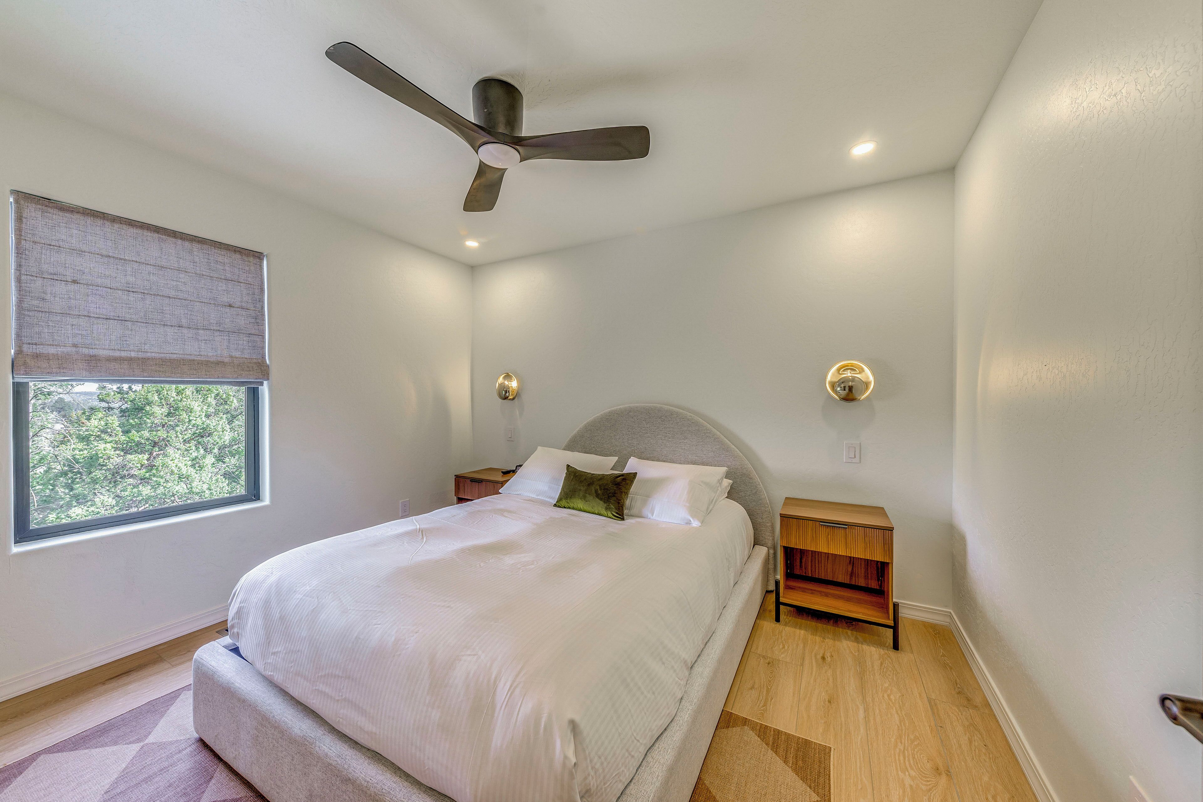 Un dormitorio con cama, ventilador de techo, mesita de noche y ventana con vista a árboles.
